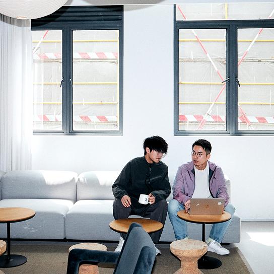 Two men sit on a light gray sofa in a bright room beneath two windows, one holding a mug while the other uses a laptop on a small round table.