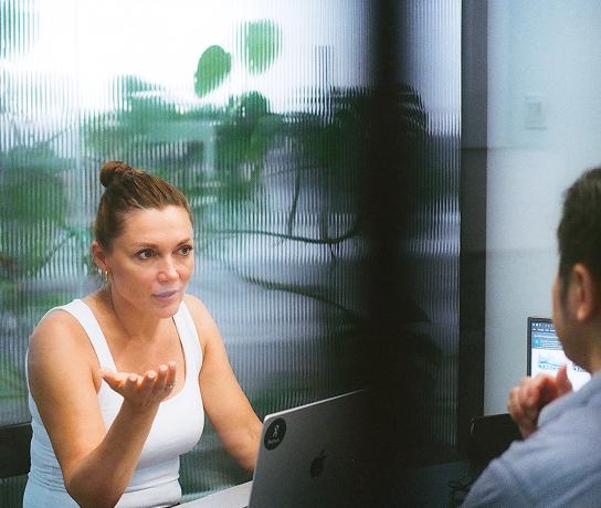 A woman in a white tank top gestures while speaking to a man across a table with a laptop in front of her, with a frosted glass panel and plants visible behind them.