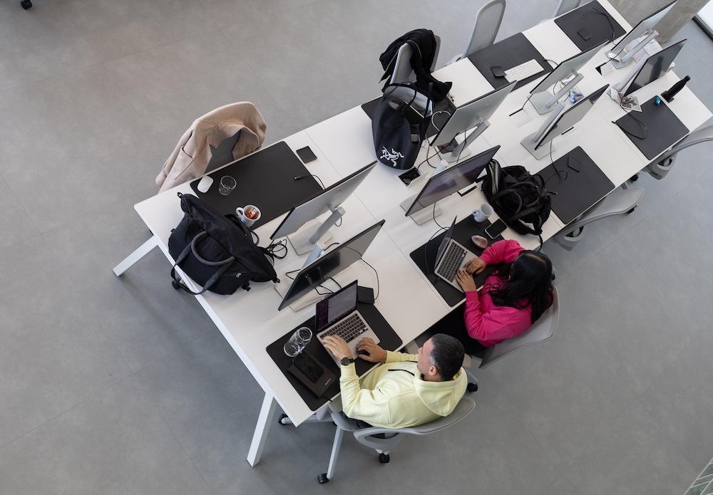 Overhead view of two people seated at a shared white office desk, each working on a laptop among monitors, backpacks, mugs, and a coat.