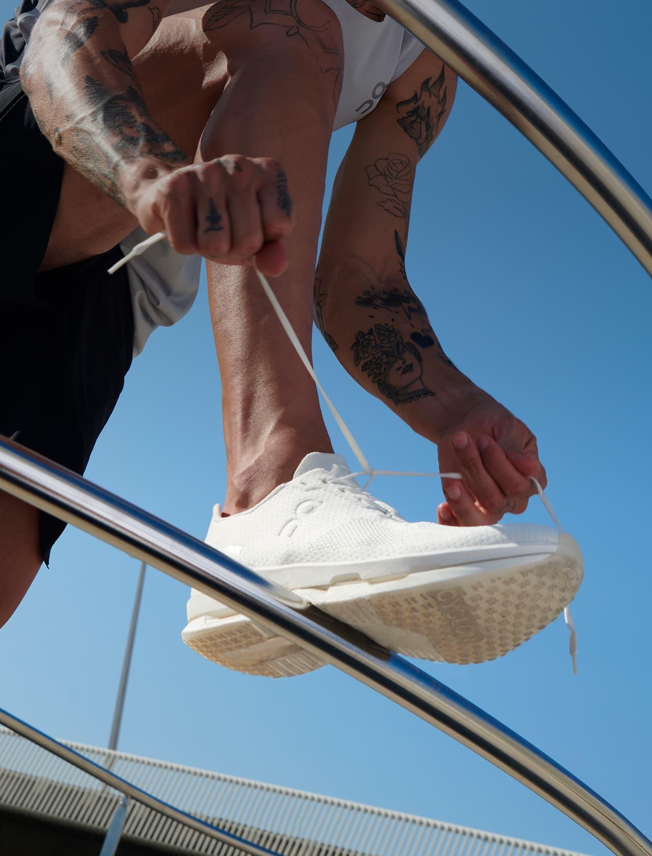 Close-up low-angle photo of a tattooed person tying the laces of a white sneaker while perched on a curved metal rail against a blue sky.