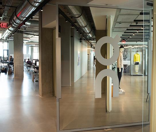 Open-plan office interior with exposed ductwork, a glass partition bearing a large stylized circular logo, workstations and people visible on the left, and a mannequin wearing a yellow jacket in a display case on the right.
