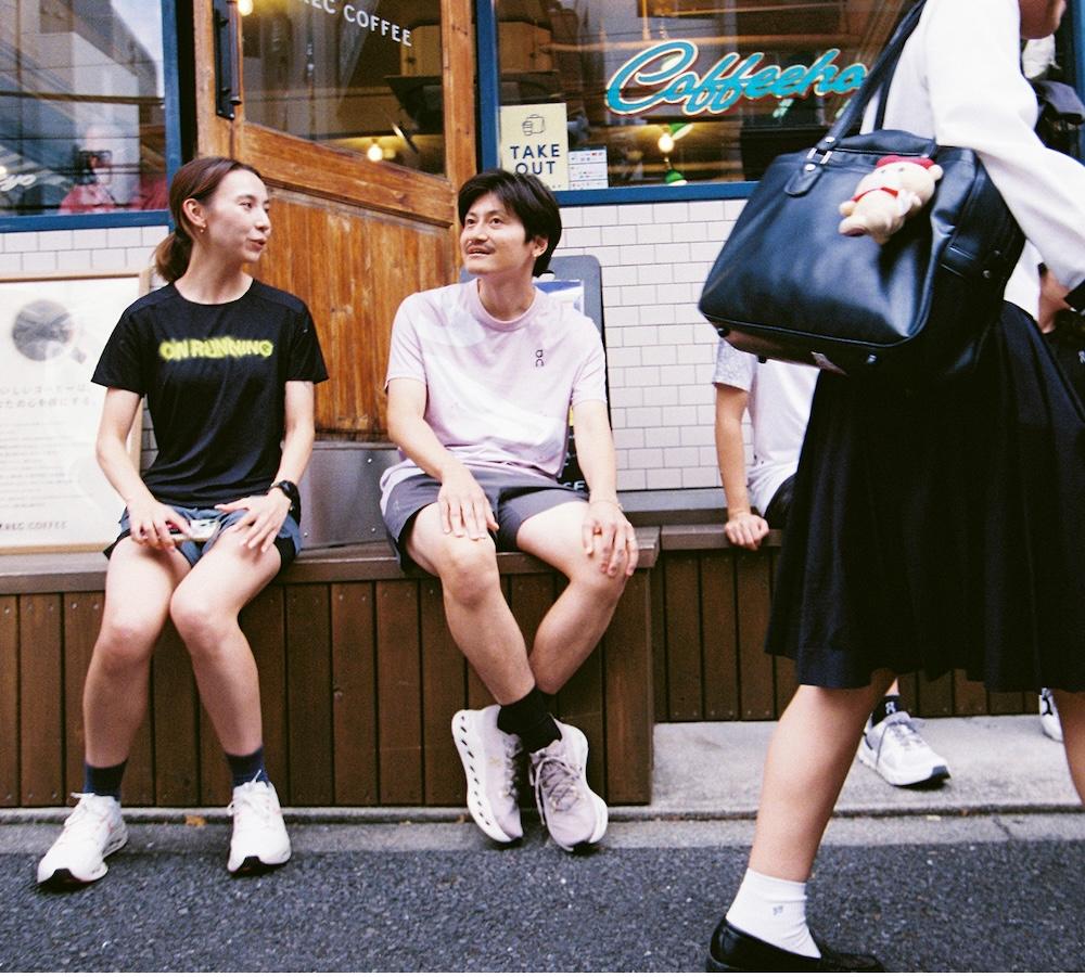 Two people sit on a wooden bench outside a coffee shop talking while another person walks past carrying a large black bag with a small plush toy attached.