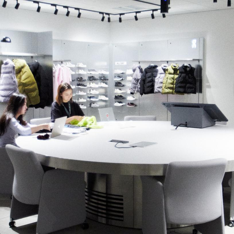 A bright room with a large round white table and chairs in the foreground and a wall display of shoes and four puffer jackets hanging above a counter in the background.