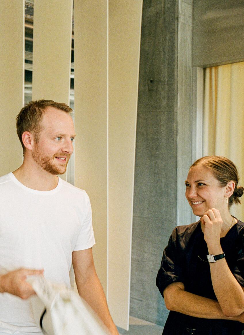 Man in a white T-shirt holding a fabric item and smiling while talking to a woman in black who is smiling with her hand near her chin, standing in a modern room with tall vertical panels.