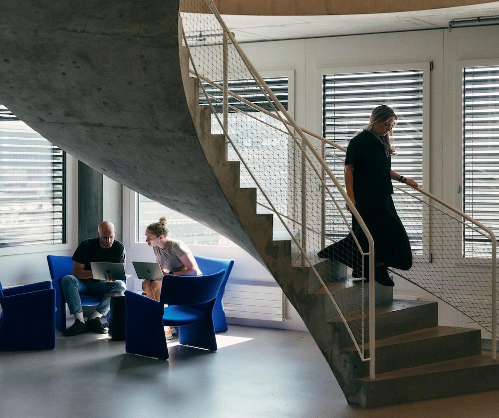 Two people sit in blue chairs working on laptops beneath a concrete staircase while a third person descends the stairs beside windows with horizontal blinds.