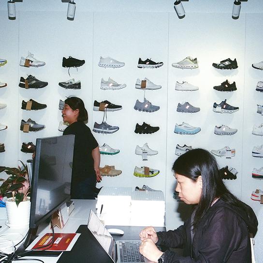 Two people in a shoe shop workspace, one seated at a desk using a laptop and the other standing in front of a white wall displaying rows of sneakers.