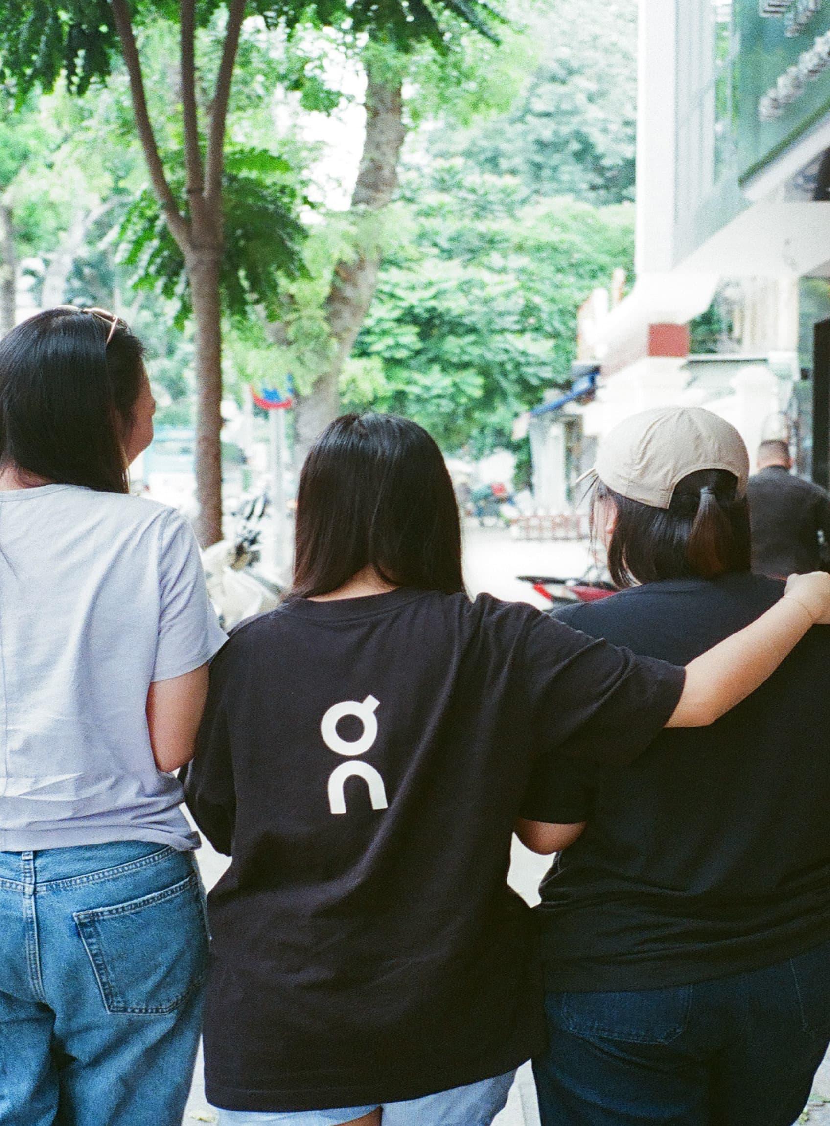 Three women walk arm in arm down a tree-lined sidewalk, seen from behind.