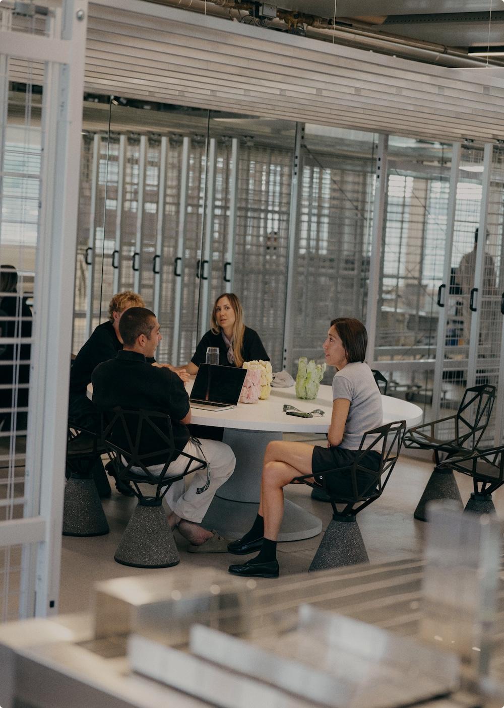 Four people sit around a round white table in a modern office meeting area, with a laptop and decorative objects on the table.