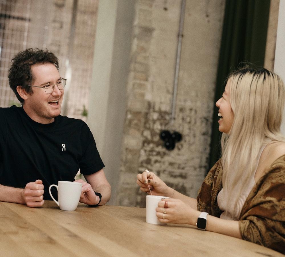 A man wearing glasses and a black t-shirt and a woman with long blonde hair sit at a wooden table, each holding a mug and laughing.
