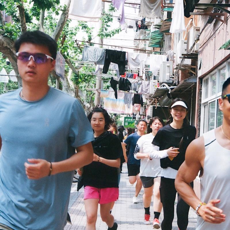 A group of people jogging down a narrow residential alley lined with hanging laundry and air-conditioning units.