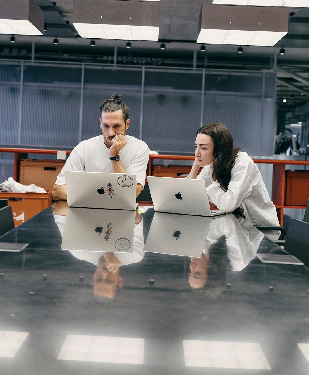 Two people sit side by side at a glossy table, each looking at a silver Apple laptop with their reflections visible on the table surface.