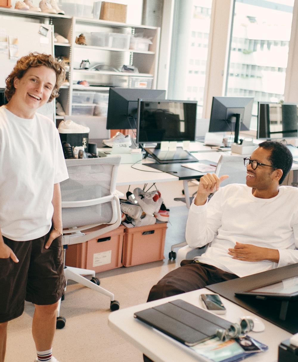 Two people in a bright office smiling and talking, one standing and one seated and gesturing, with computer monitors and storage boxes behind them.