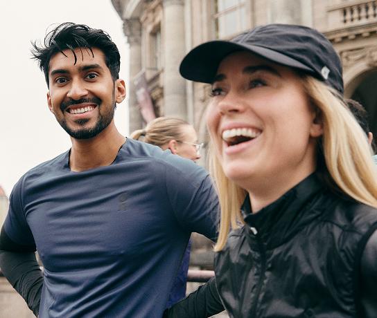 A smiling man in a blue shirt stands beside a laughing woman wearing a black cap and jacket in front of a city building.