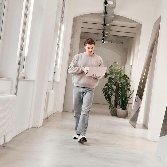 A man in a sweatshirt and jeans walks down a bright hallway while holding and looking at a laptop.