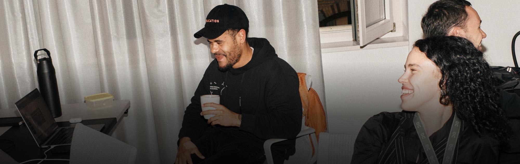 Two people laugh together during a casual meeting, one holding a mug near a laptop and water bottle.