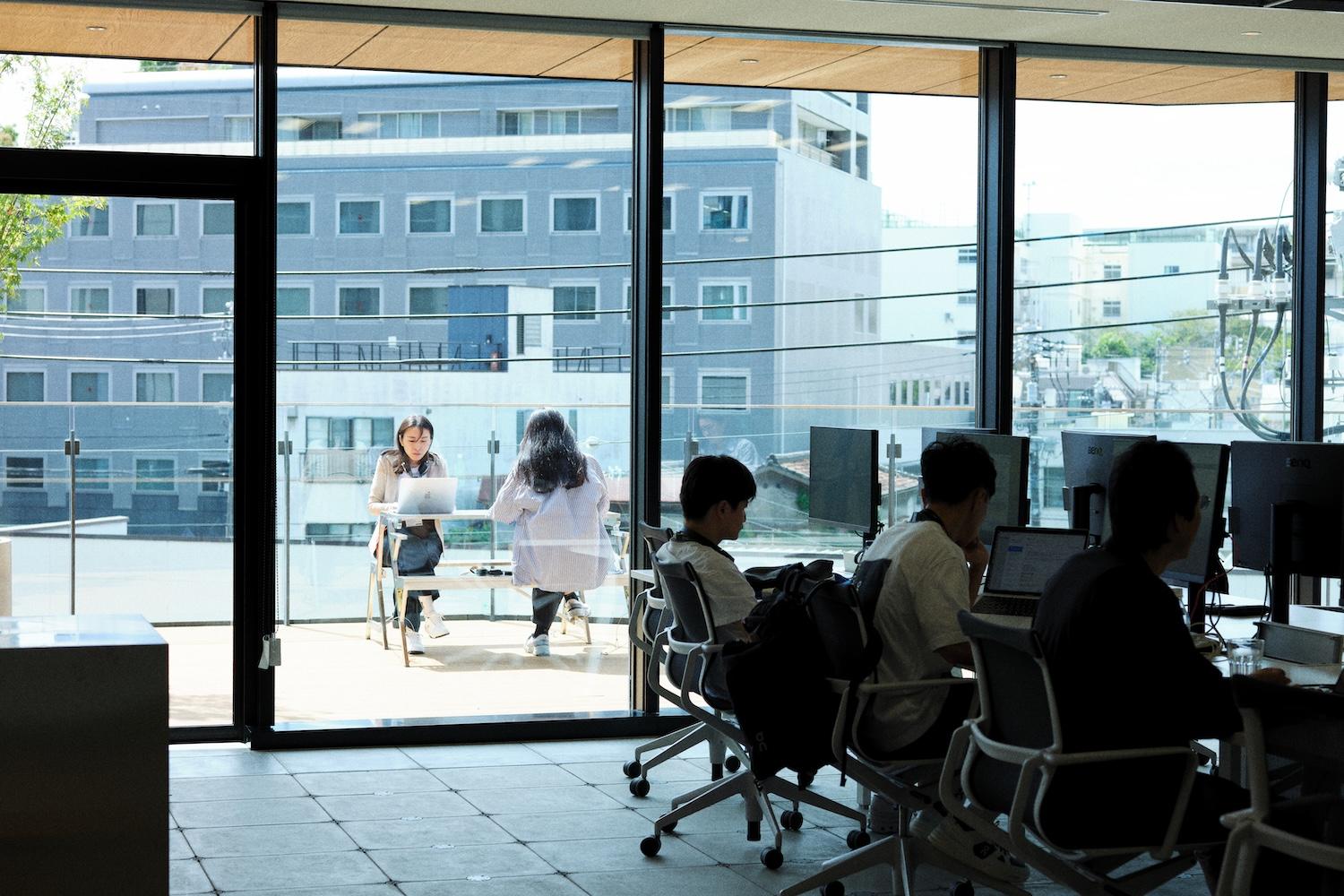 Indoor and outdoor workspaces blend as two women sit on a terrace with laptops while several colleagues work at desks inside.
