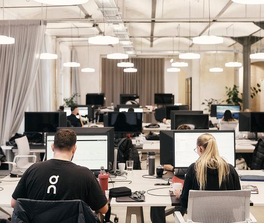 Open-plan office with rows of desks and computer monitors, two people seated in the foreground working at their screens.
