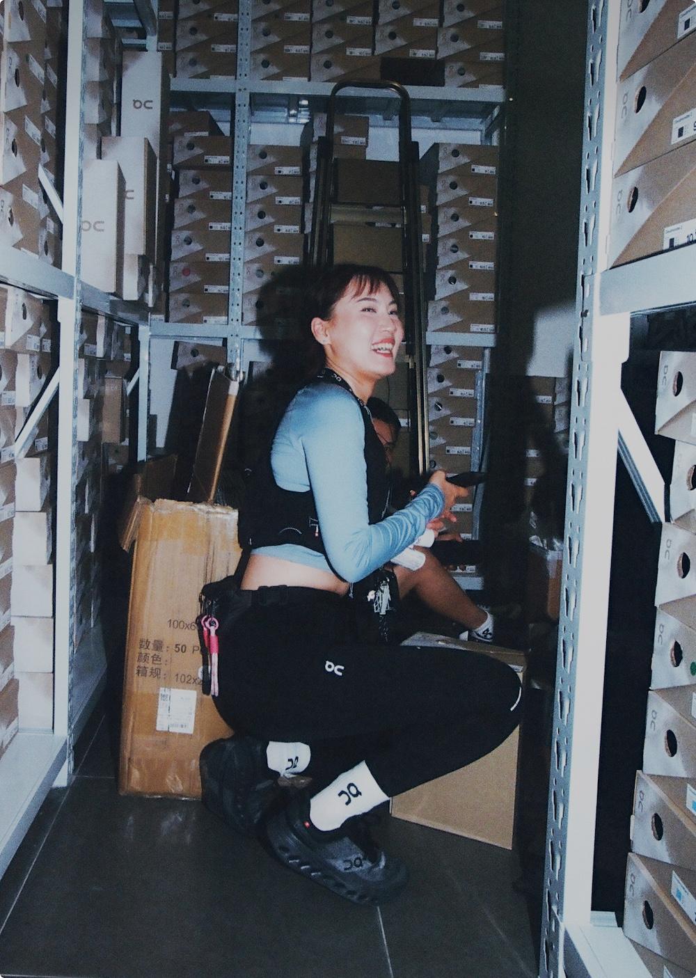 A smiling woman crouches between metal shelving units stacked with shoeboxes in a storage room.