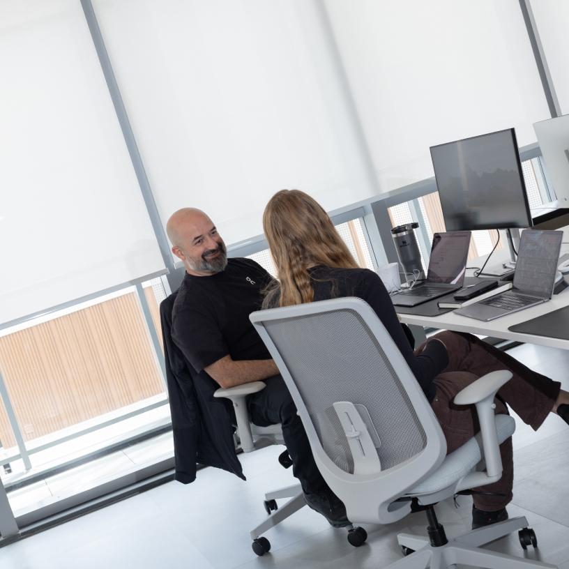Two coworkers sit in office chairs at adjacent desks—one bald man smiling and facing a long-haired colleague—surrounded by laptops and monitors.