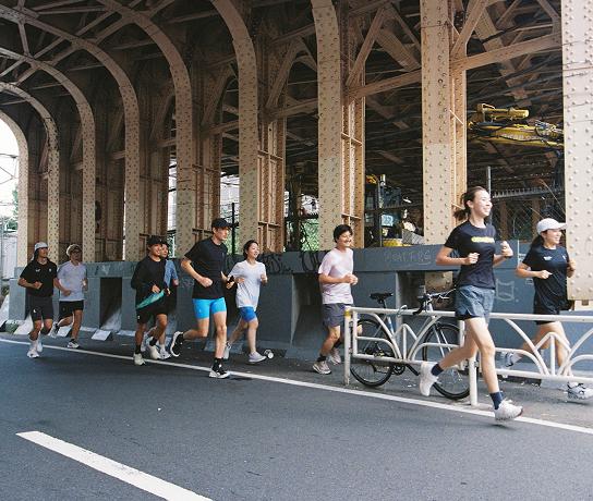 A group of people jogging along a city street beneath a riveted steel overpass, with a bicycle parked against a railing.