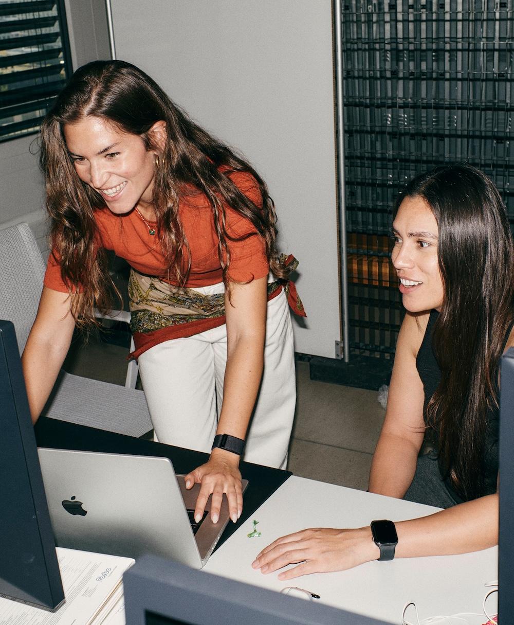 Two people at a desk smiling and looking at a laptop, with one leaning over the keyboard while the other sits beside them.