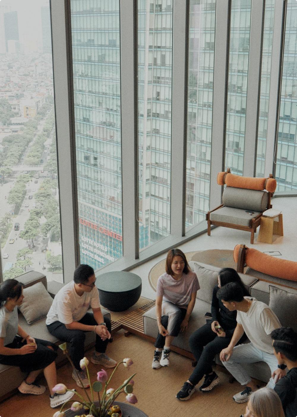 Several people sit and converse in a modern high-rise lounge with floor-to-ceiling windows overlooking a cityscape.