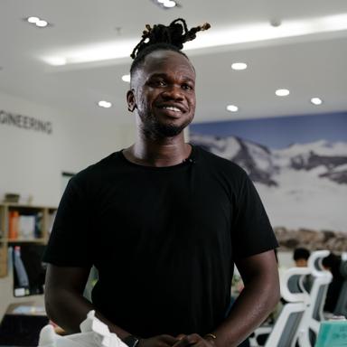 A man in a black t-shirt with his hair tied up is standing and smiling