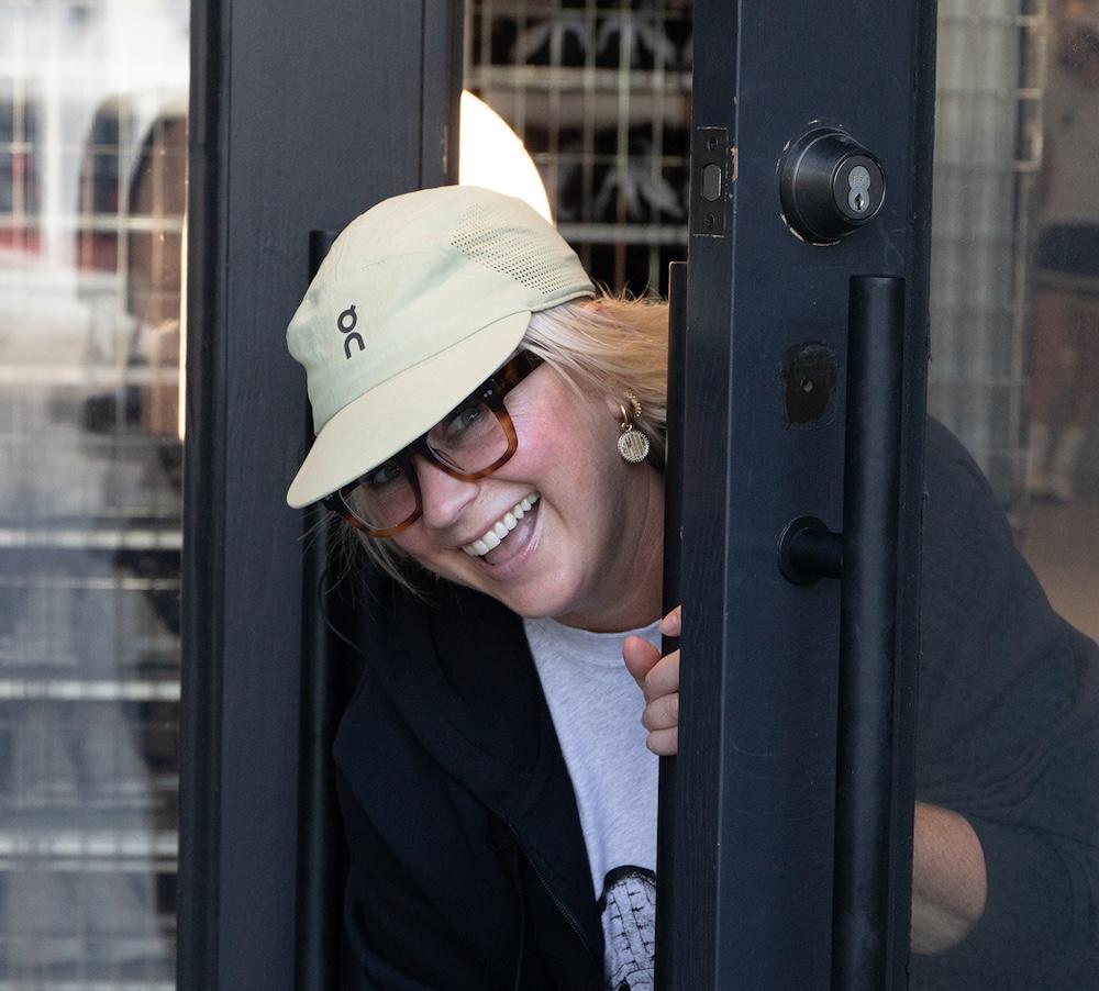 Person wearing a light green cap, glasses, and earrings smiles while peeking from behind a partially open dark door and holding its handle.