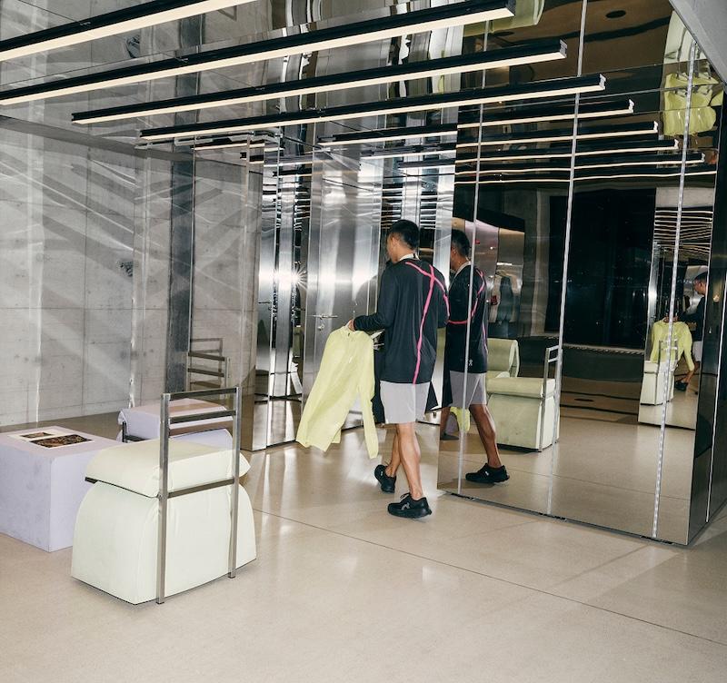 Man holding a yellow shirt walks through a modern room with mirrored walls, pastel furniture, and ceiling light beams.
