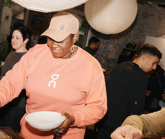A smiling person in a peach sweatshirt and matching cap holds a white bowl while serving food at an indoor gathering beneath hanging paper lanterns.