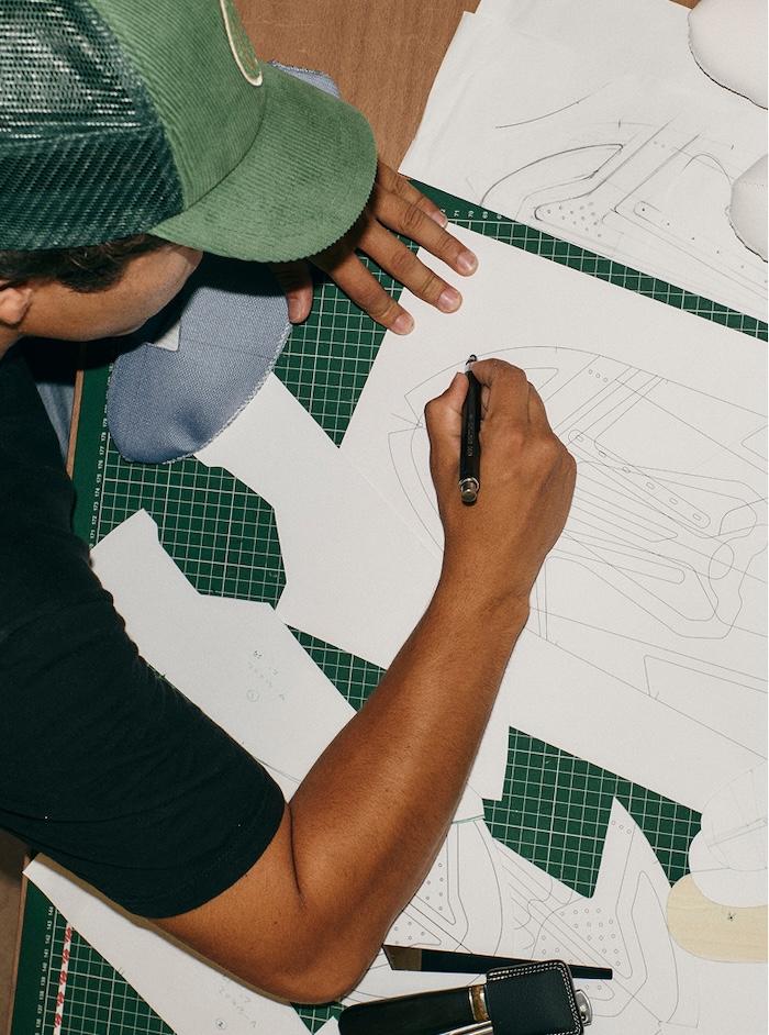 A person wearing a green cap sketches shoe designs on paper atop a green cutting mat, surrounded by paper patterns and drawing tools.