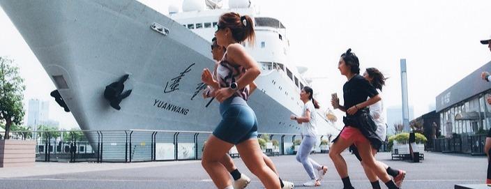 Several runners in athletic clothing jogging along a waterfront promenade past a large docked ship named "YUANWANG".