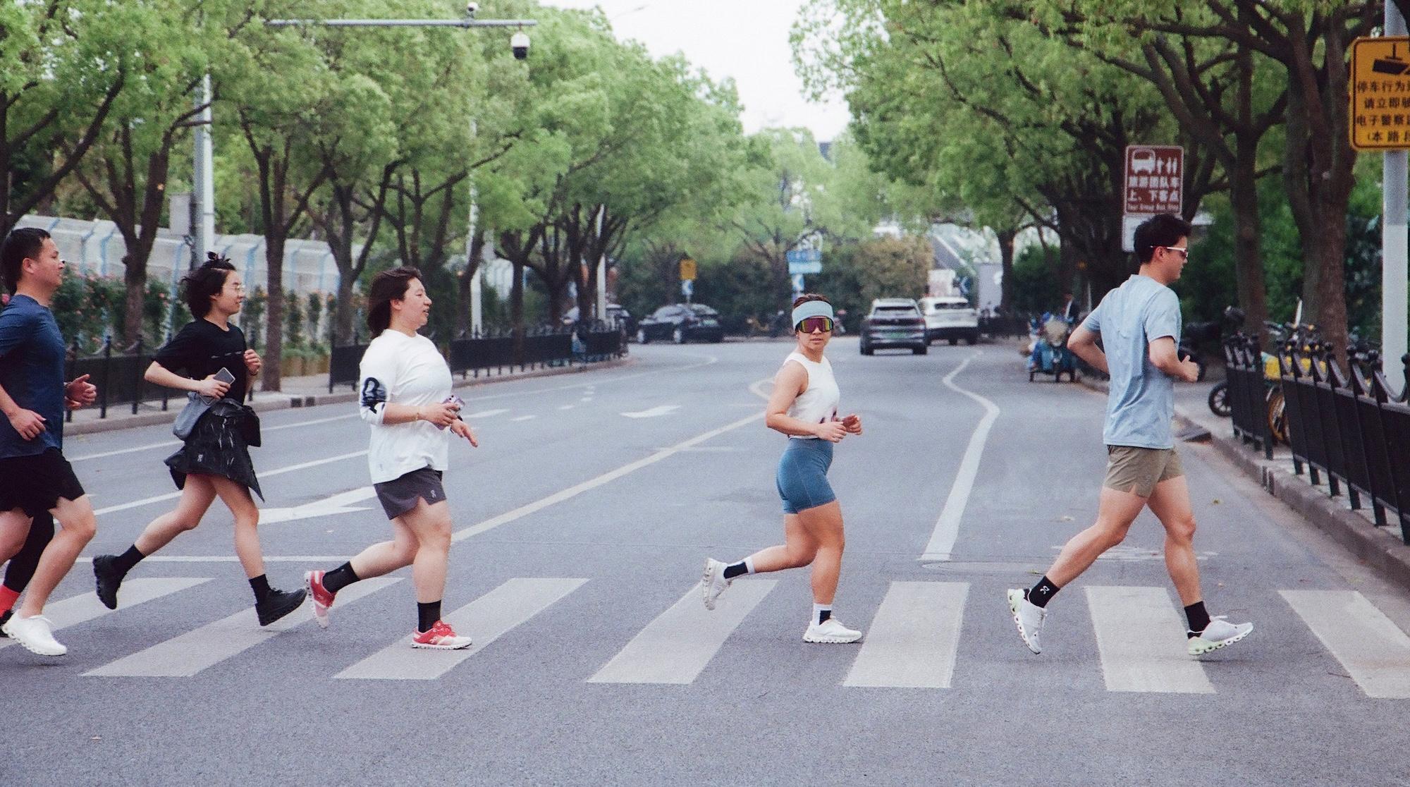 Five people jogging across a tree-lined street on a zebra crosswalk, with one woman in a visor and sunglasses looking back.