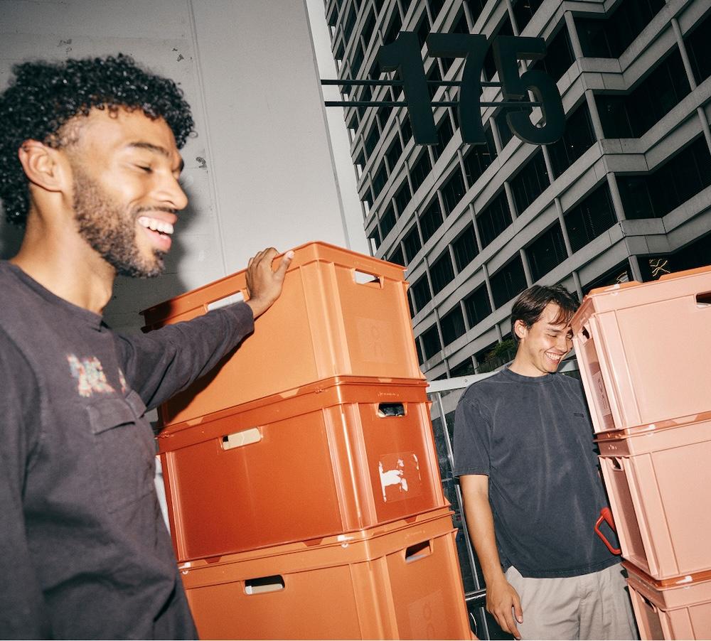 Two men smiling beside stacks of orange plastic crates with a multi-story office building marked "115" behind them.