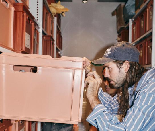 Man in a striped shirt and cap labels a pink storage bin in a narrow room lined with stacked plastic containers.