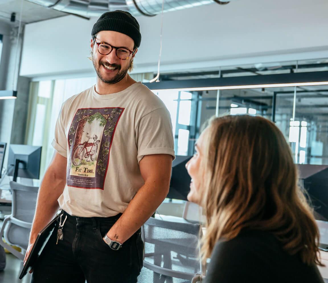 Man in a graphic T-shirt and beanie smiling while talking to a seated woman with long hair in a modern, open-plan office with bright lighting.
