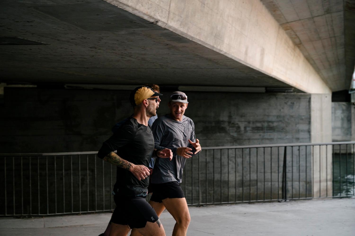 Two men in athletic gear jog under a concrete bridge, smiling and talking, with a metal railing and water in the background.