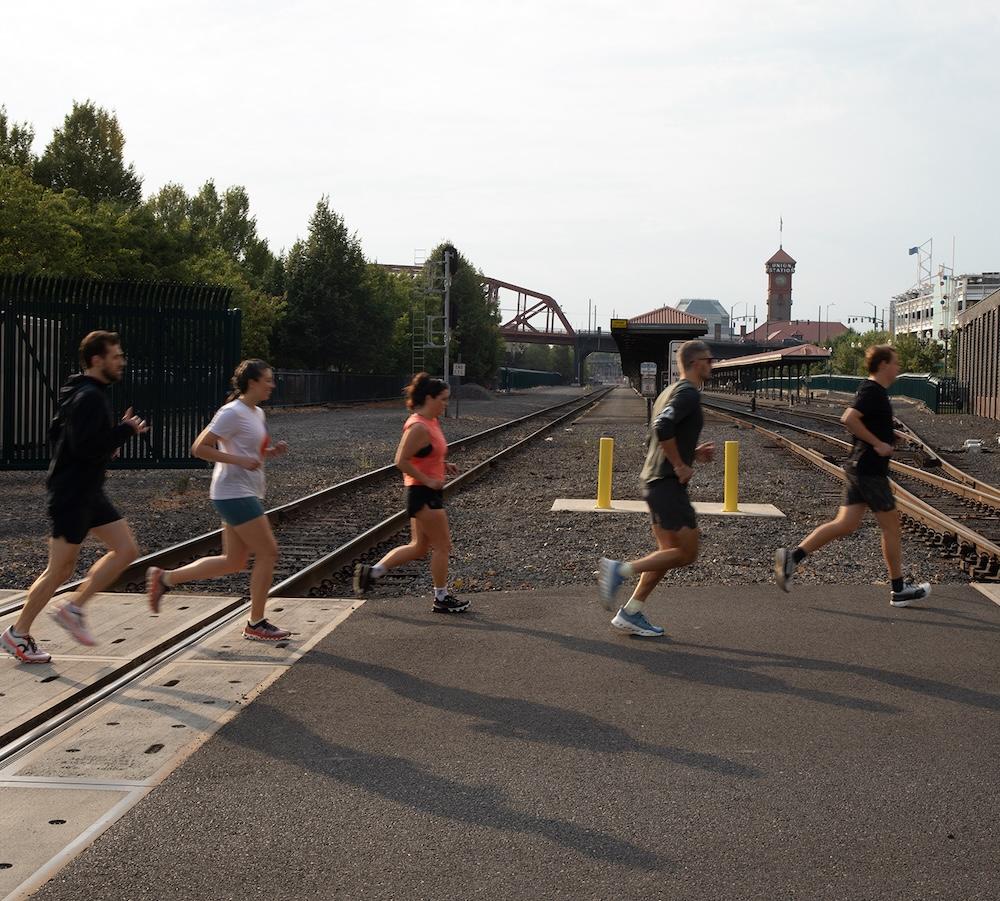 Five people jog across a railroad crossing in front of train tracks and a station platform with a clock tower in the background.