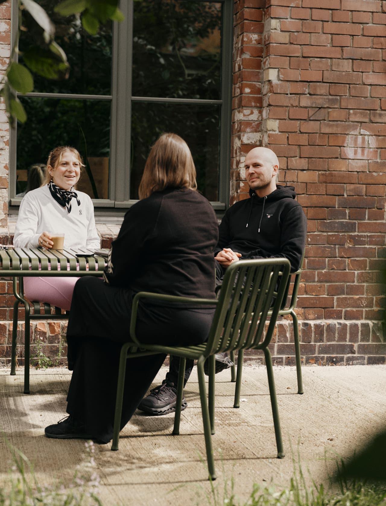 Three people sit and talk at a green metal table in a sunny courtyard beside a red brick building.