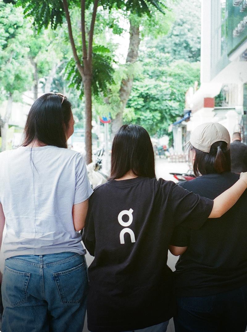 Three women walk outdoors arm-in-arm on a tree-lined street, viewed from behind.