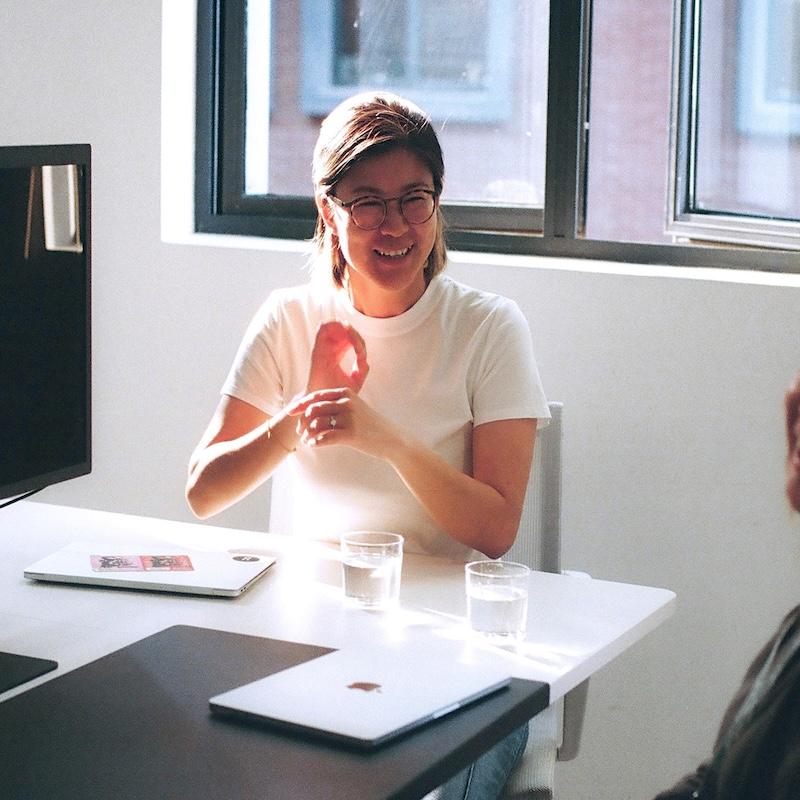 Person in a white T-shirt and glasses smiling and gesturing while seated at a table with two laptops and two glasses of water by a window.