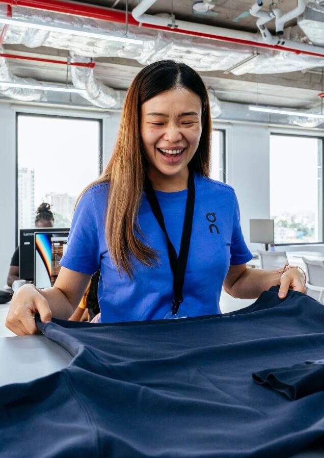 Smiling woman in a blue shirt holding a dark garment across a table in a bright, modern office with large windows and exposed ceiling pipes.