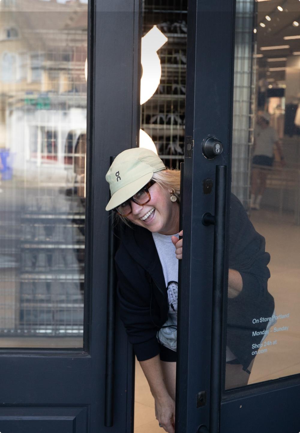 A person wearing a light-colored cap and glasses smiles while leaning through a partially open dark storefront door.