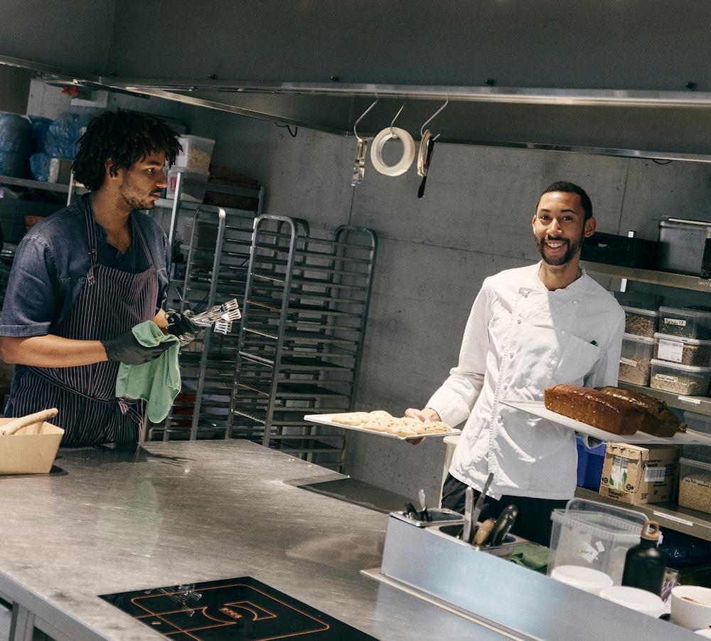 Two bakers in a commercial kitchen, one in a white chef's coat smiling while holding a tray with a loaf and a plate of cookies, and the other in a striped apron holding spatulas and a green cloth beside metal baking racks.