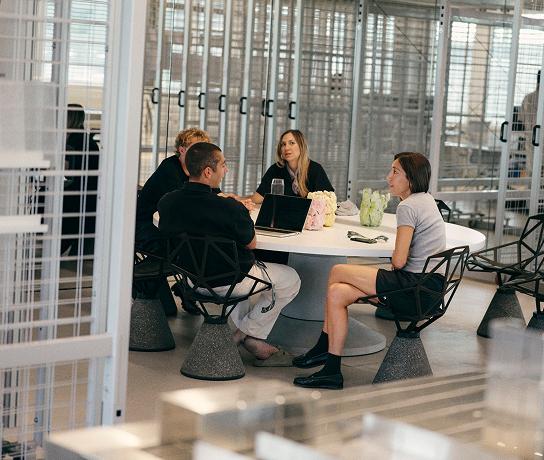 Four people sit around a round table with a laptop, notebooks, and small vases in a modern glass-walled office.