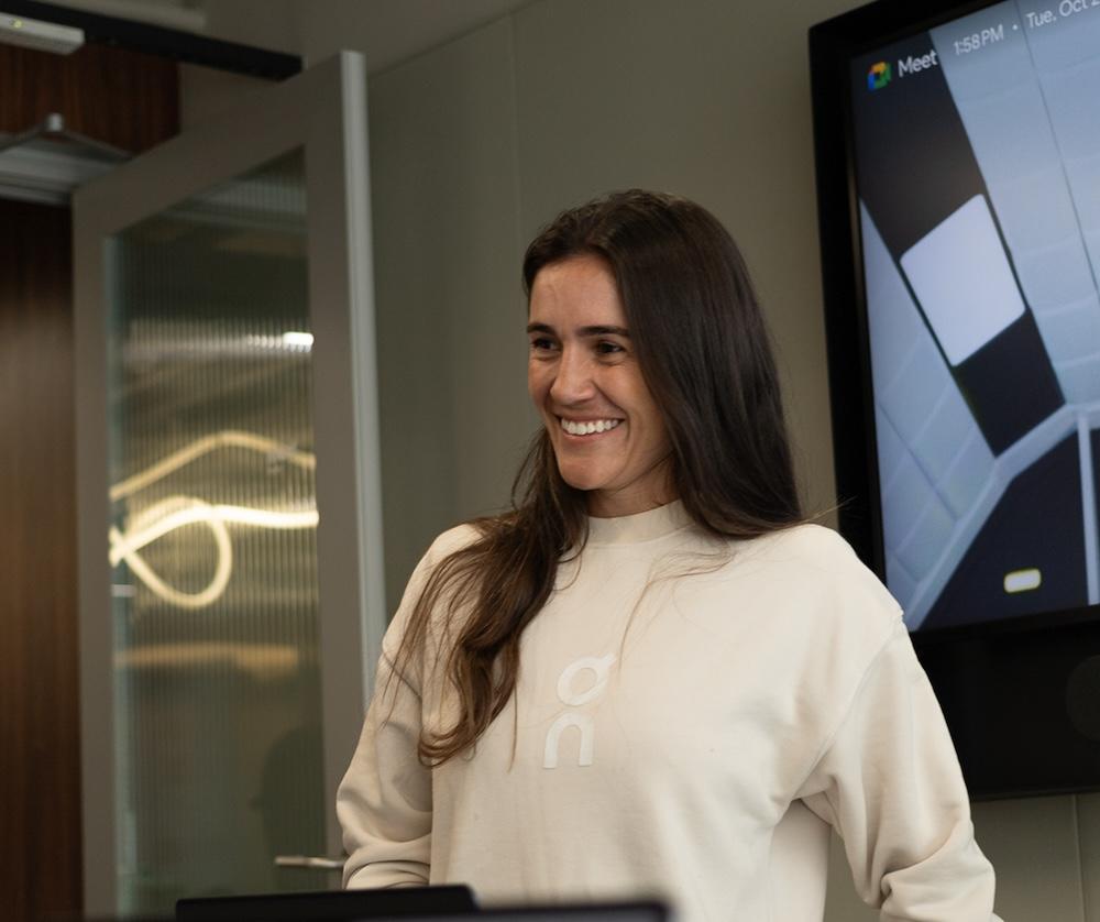A smiling woman in a light-colored sweatshirt stands in a conference room with a glass door and a large wall-mounted monitor behind her.