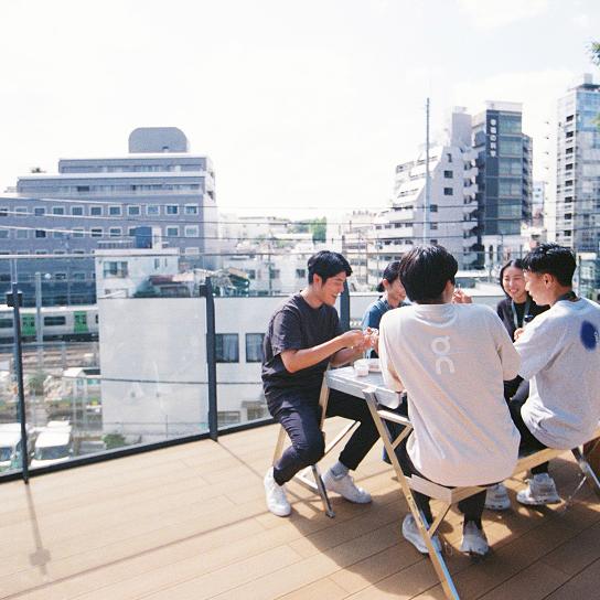 A group of people sit around a small table on a rooftop terrace overlooking city buildings behind a glass railing.