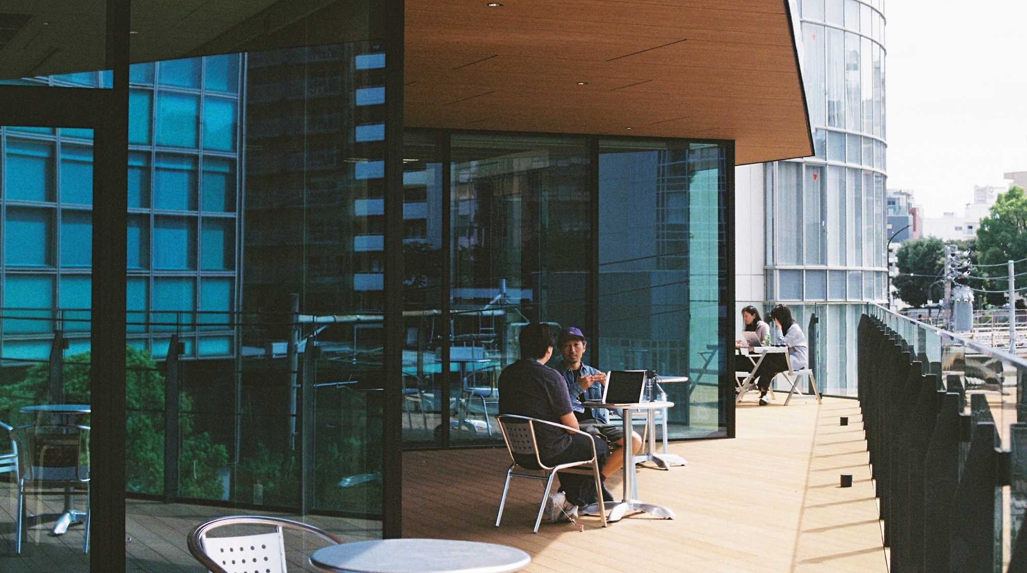 Two people sit at a small round table with a laptop on a wooden outdoor terrace beside a modern glass building, while two others sit at a table further down the deck.