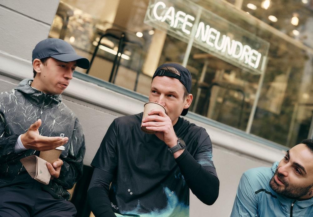 Three men in athletic clothing sit outside a cafe with a neon "CAFE NEUNDREI" sign, one drinking from a paper cup while another gestures and the third listens.