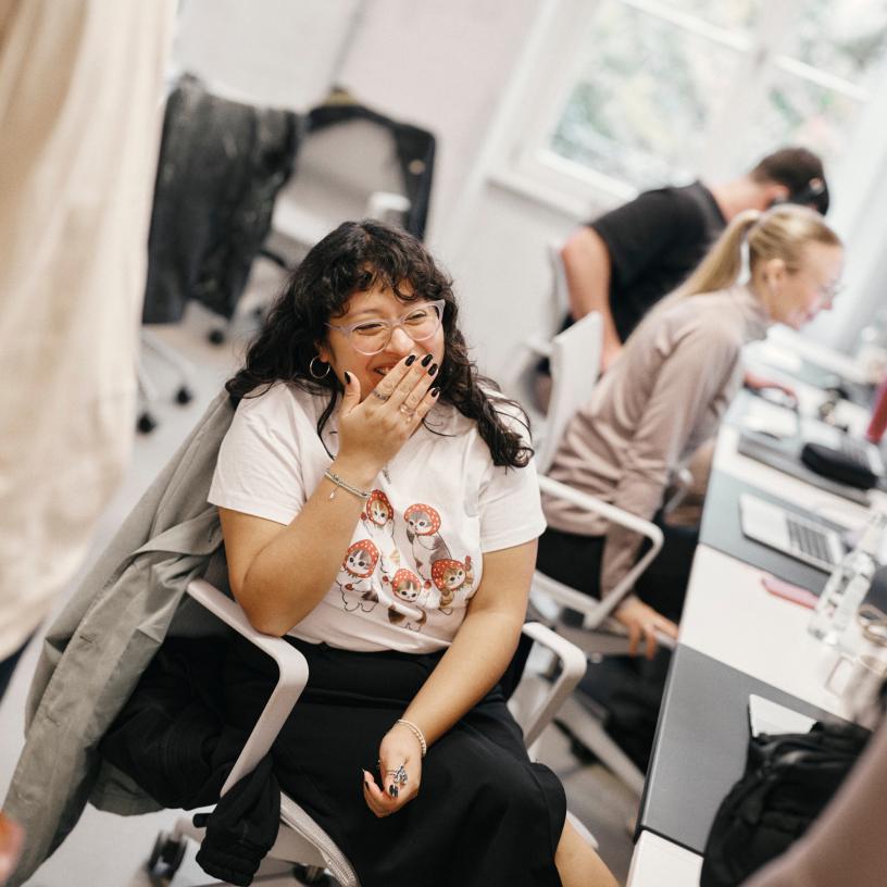 A person wearing glasses and a cartoon t-shirt laughs with a hand over their mouth while two colleagues work at desks with laptops in the background.